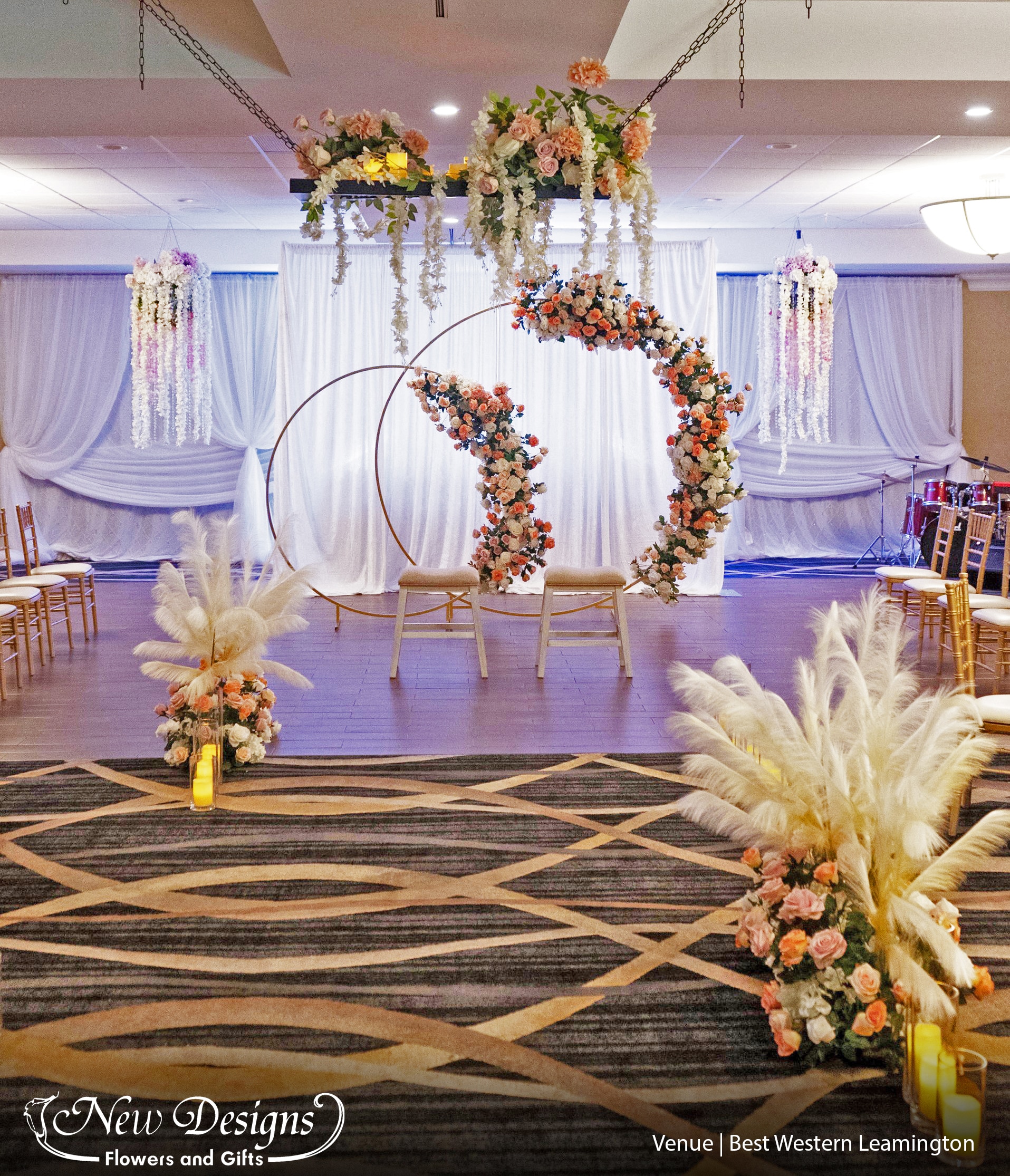 A double Circle Arch accented with Spiking Pampas grass, and installments of roses in white cream and orange. Staggering floral arrangements and pillar vases with floating candles line the aisleway, and a hanging ceiling installment of the same flowers accent the room.